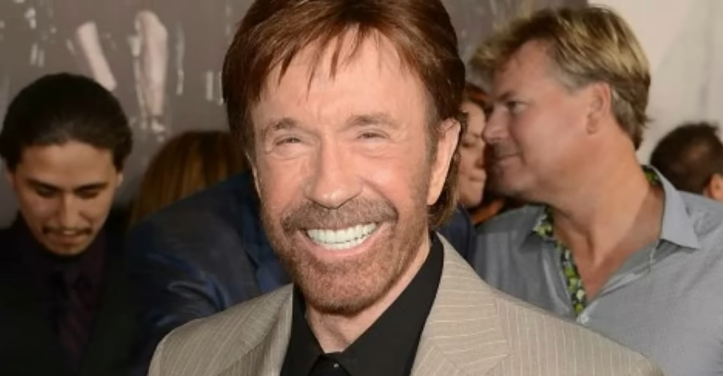 Chuck Norris in a beige jacket and black shirt standing and smiling among other people behind